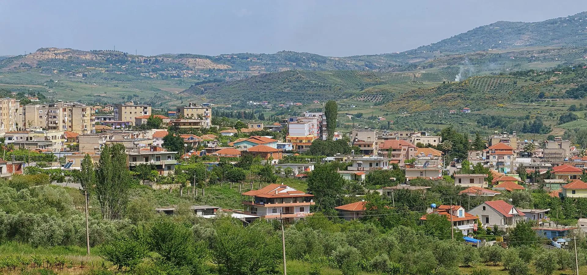 Panorámica de Ballsh, con casas de tejados rojos y bloques de apartamentos entre olivares al pie de verdes colinas