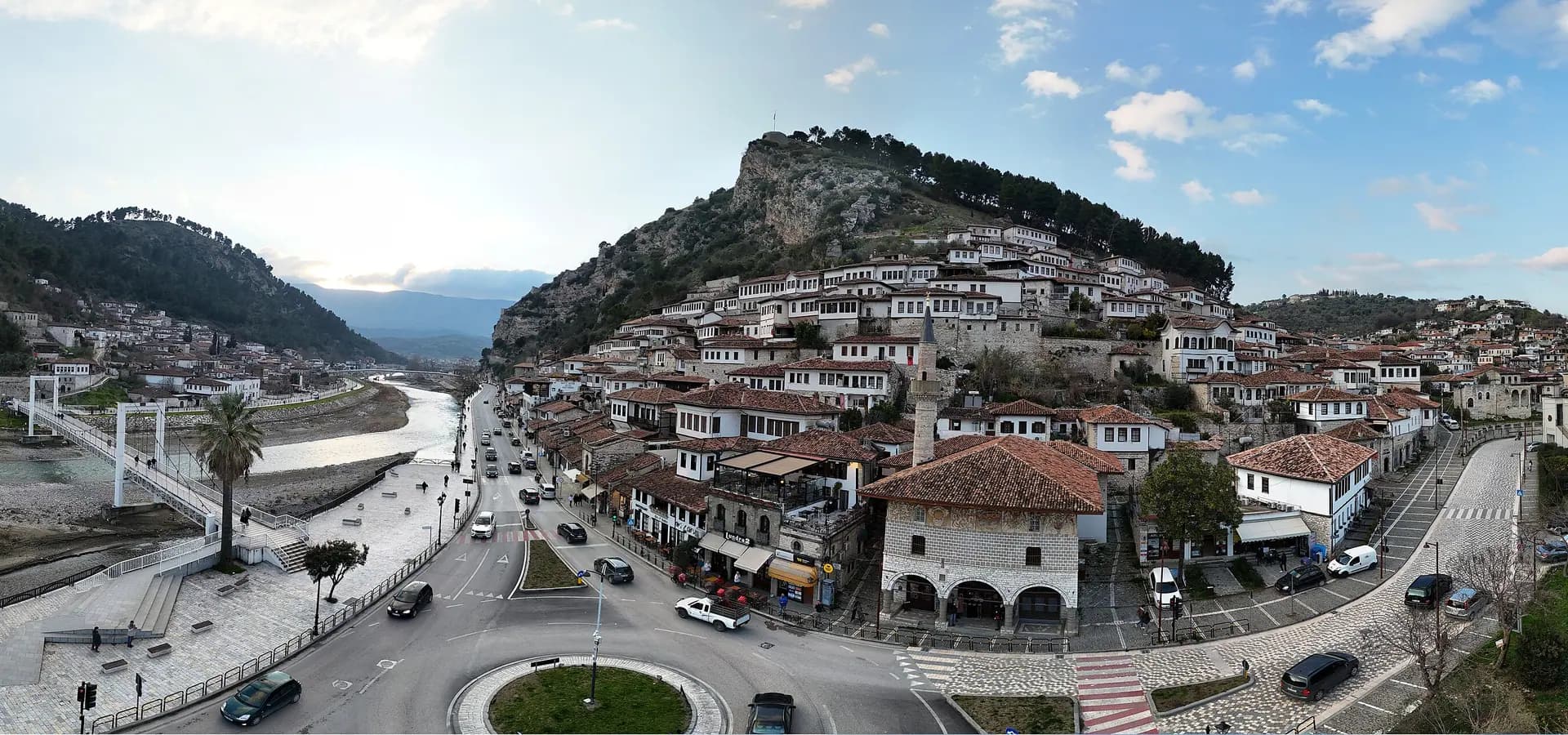 Panorama de Berat com o rio Osum, uma passarela e casas brancas subindo rumo à colina do castelo