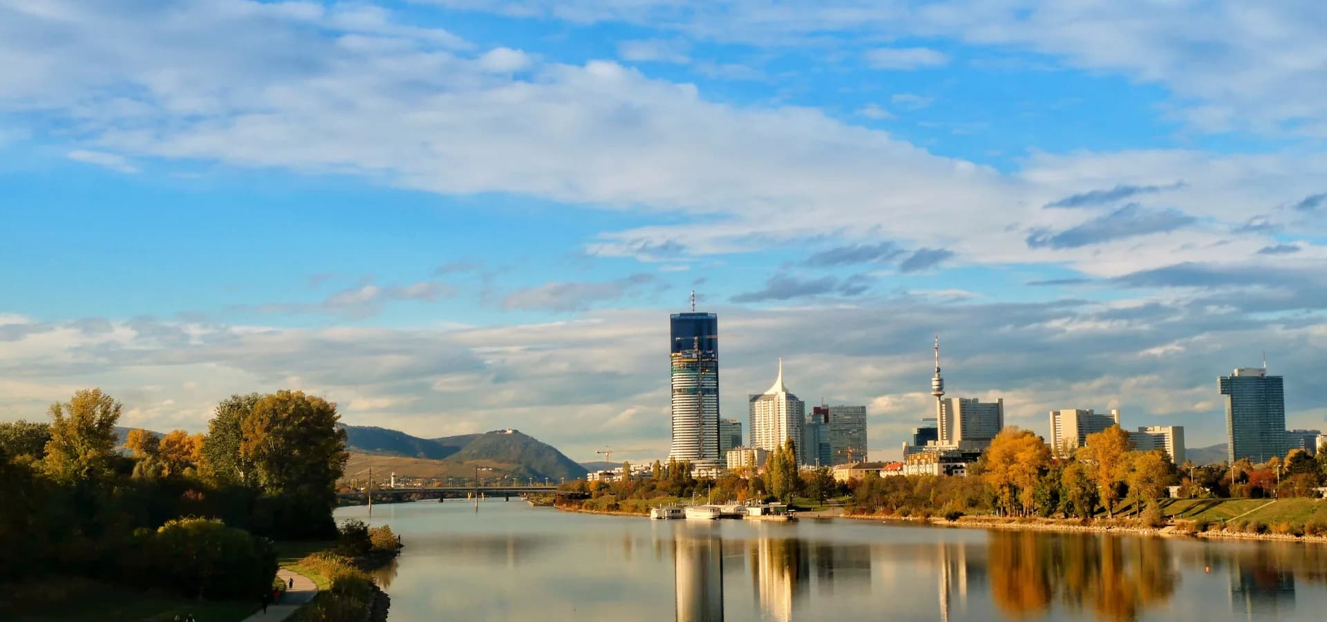 Vienna's Danube skyline in autumn, towers reflected in calm water under a wide blue sky with soft clouds
