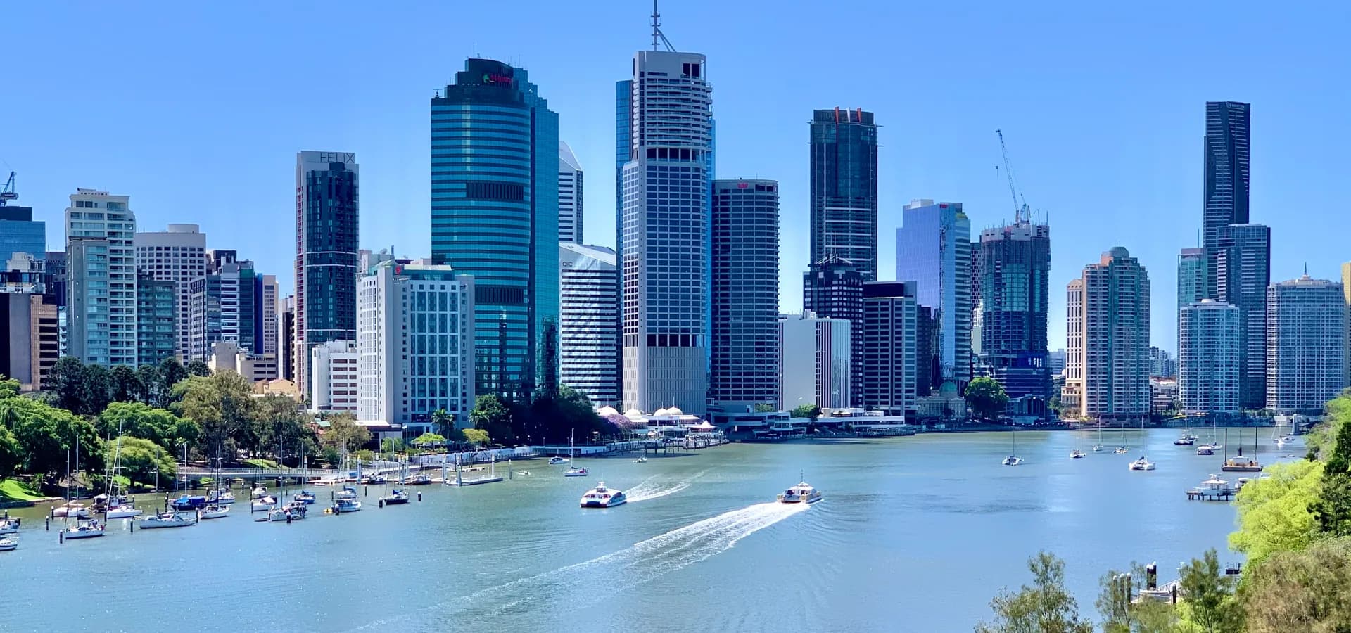 Glastürme von Brisbane über einer weiten Flussbiegung, Fähren ziehen weiße Spuren unter klarem blauem Himmel