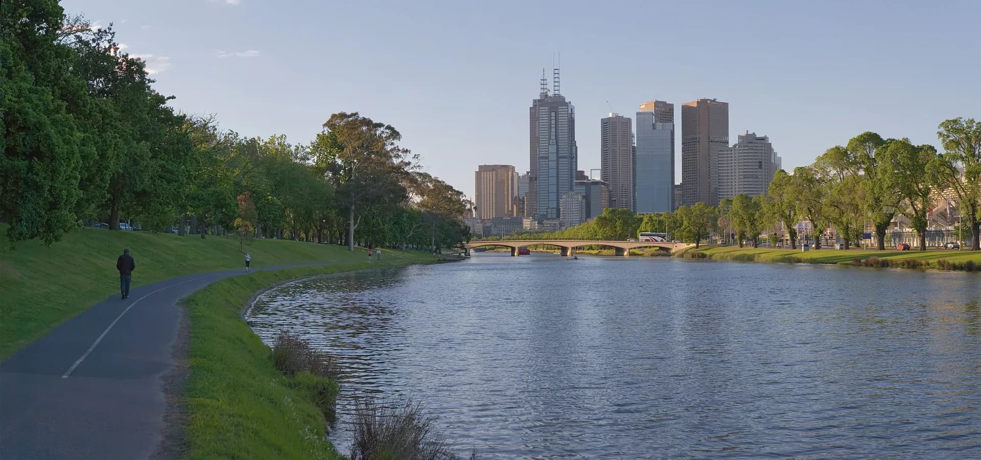 Melbourne skyline rising beyond the calm Yarra River, with tree-lined riverbanks and a walking path