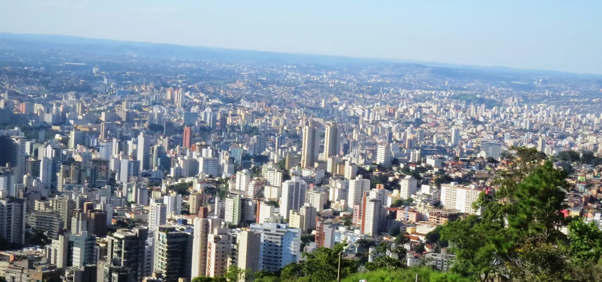 Edificios de Belo Horizonte extendiéndose sobre colinas verdes hasta un horizonte brumoso bajo un cielo azul despejado