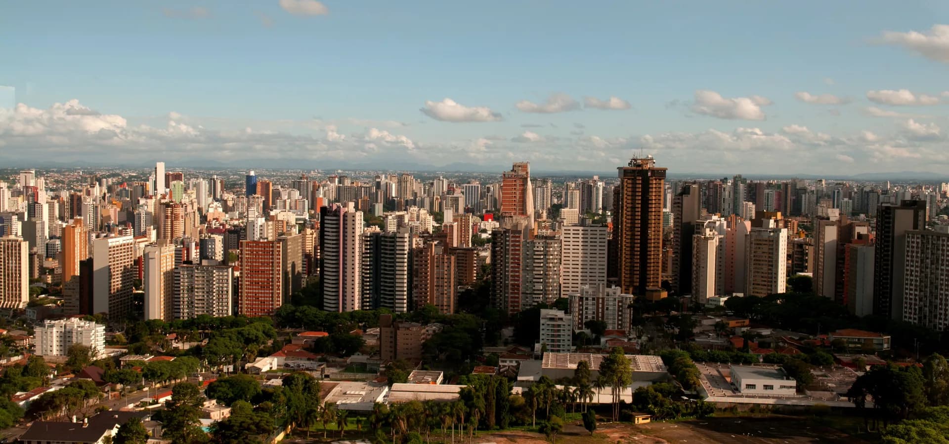 Curitiba's high-rise skyline stretching to the horizon under a wide blue sky, with green trees in the foreground