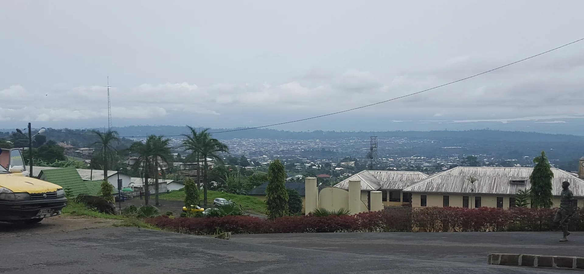 Buea se estendendo abaixo da Governor's Street, palmeiras e telhados de zinco sob um céu nublado e suave, com colinas ao fundo