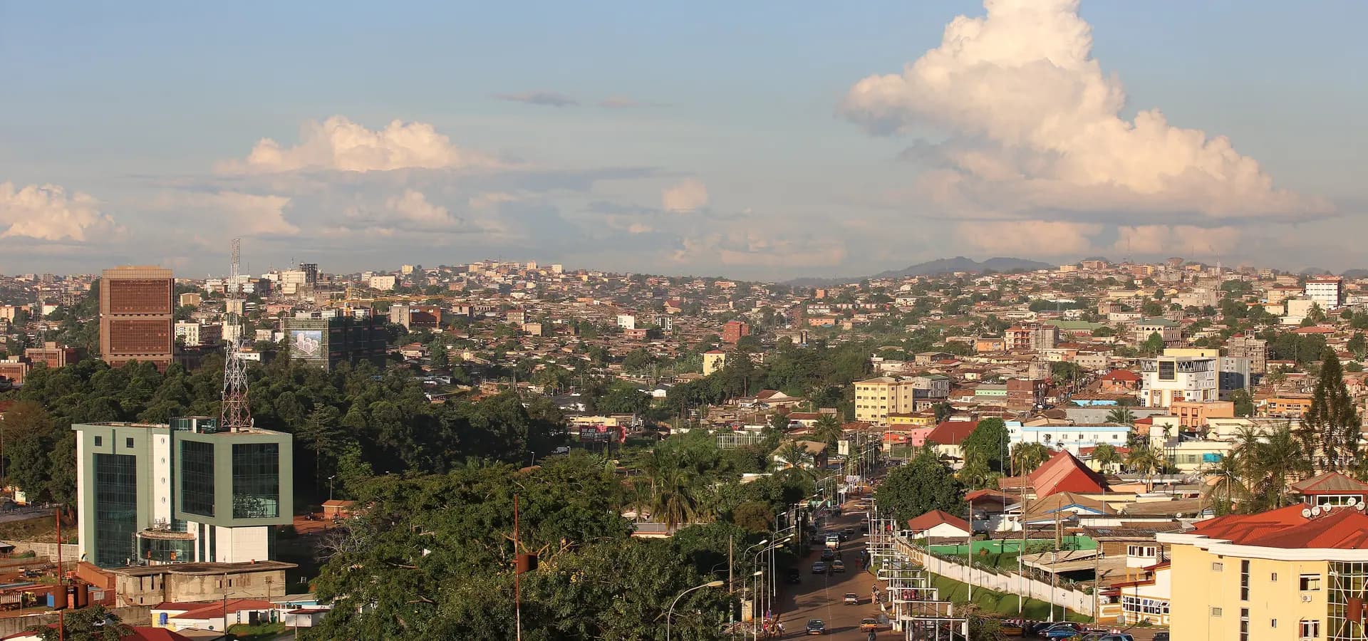 Yaoundé spreading over green hills in warm evening light, a tree-lined avenue running toward rooftops and distant peaks