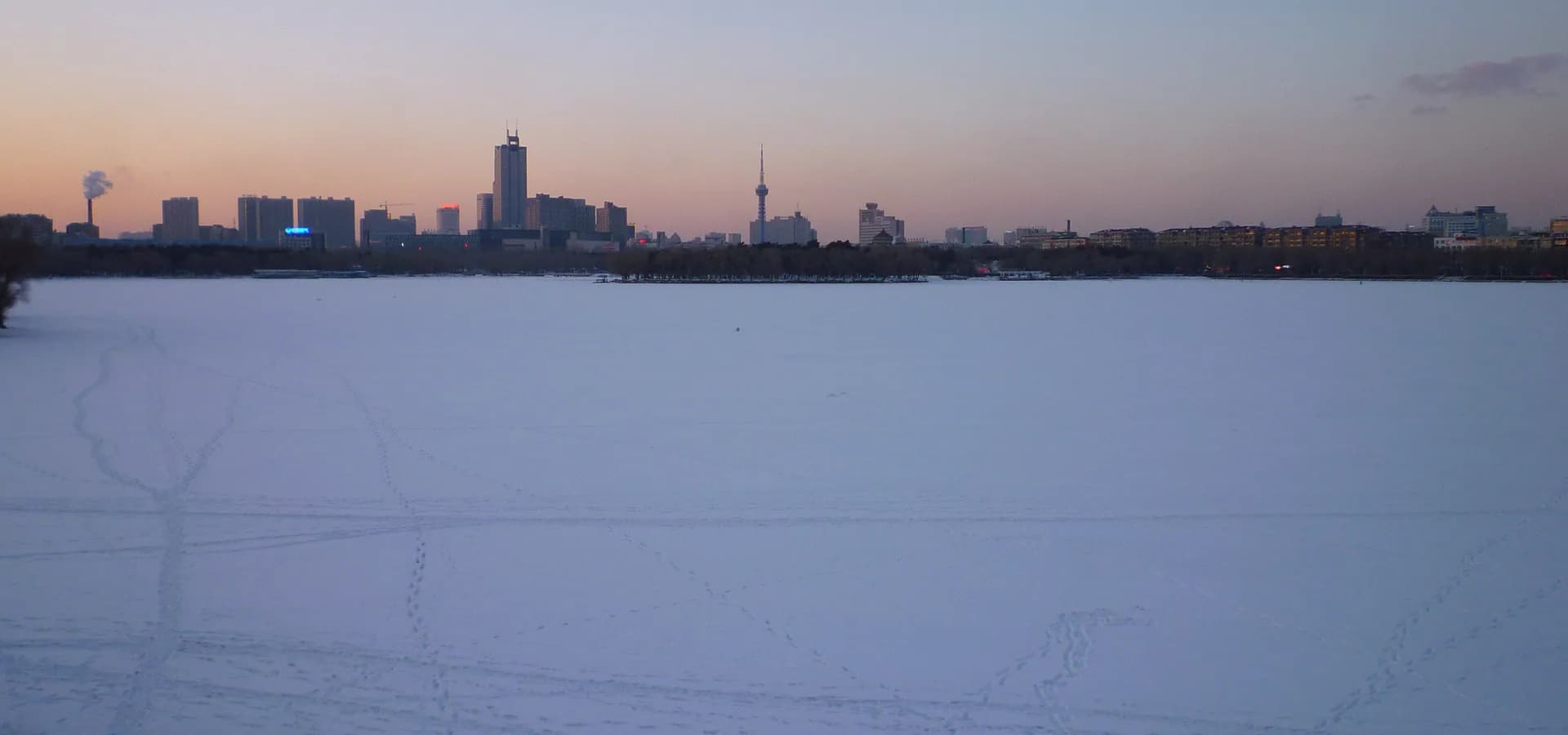 Lago Nanhu congelado e coberto de neve em Changchun, com o horizonte da cidade e a torre de TV sob um céu rosado de entardecer