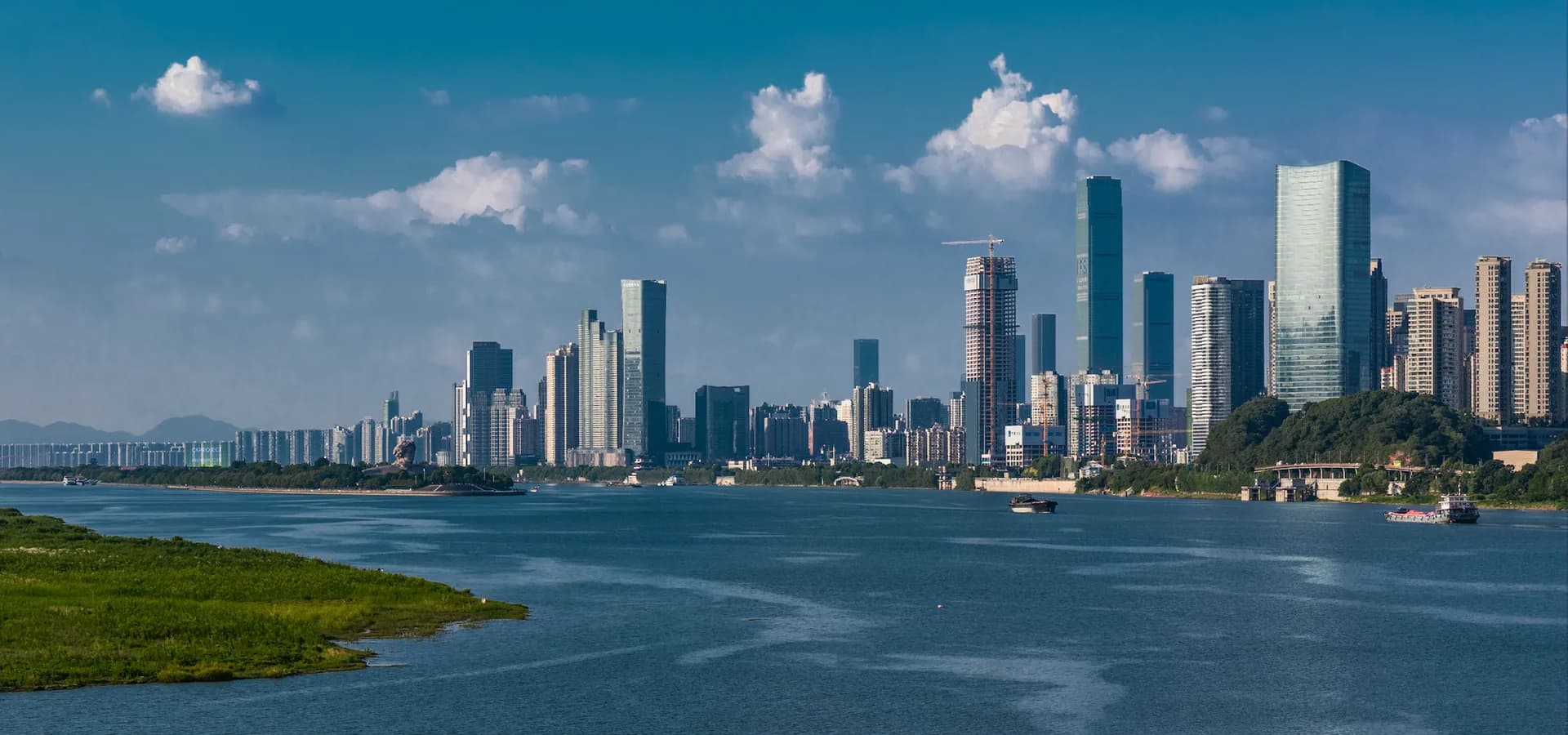 Changsha high-rises along the wide Xiang River under a blue sky, with a grassy riverbank in the foreground