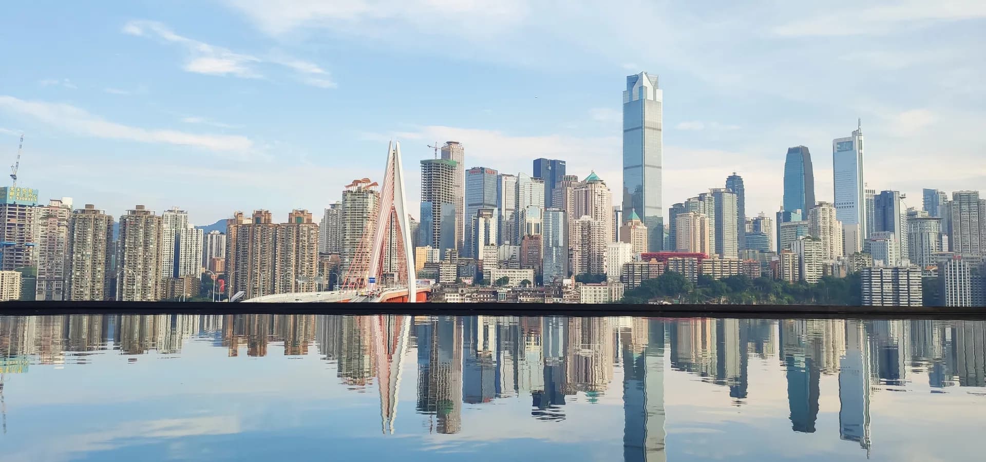 Skyline der Halbinsel Yuzhong in Chongqing mit der Qiansimen-Brücke, gespiegelt im ruhigen Wasser unter blassblauem Himmel