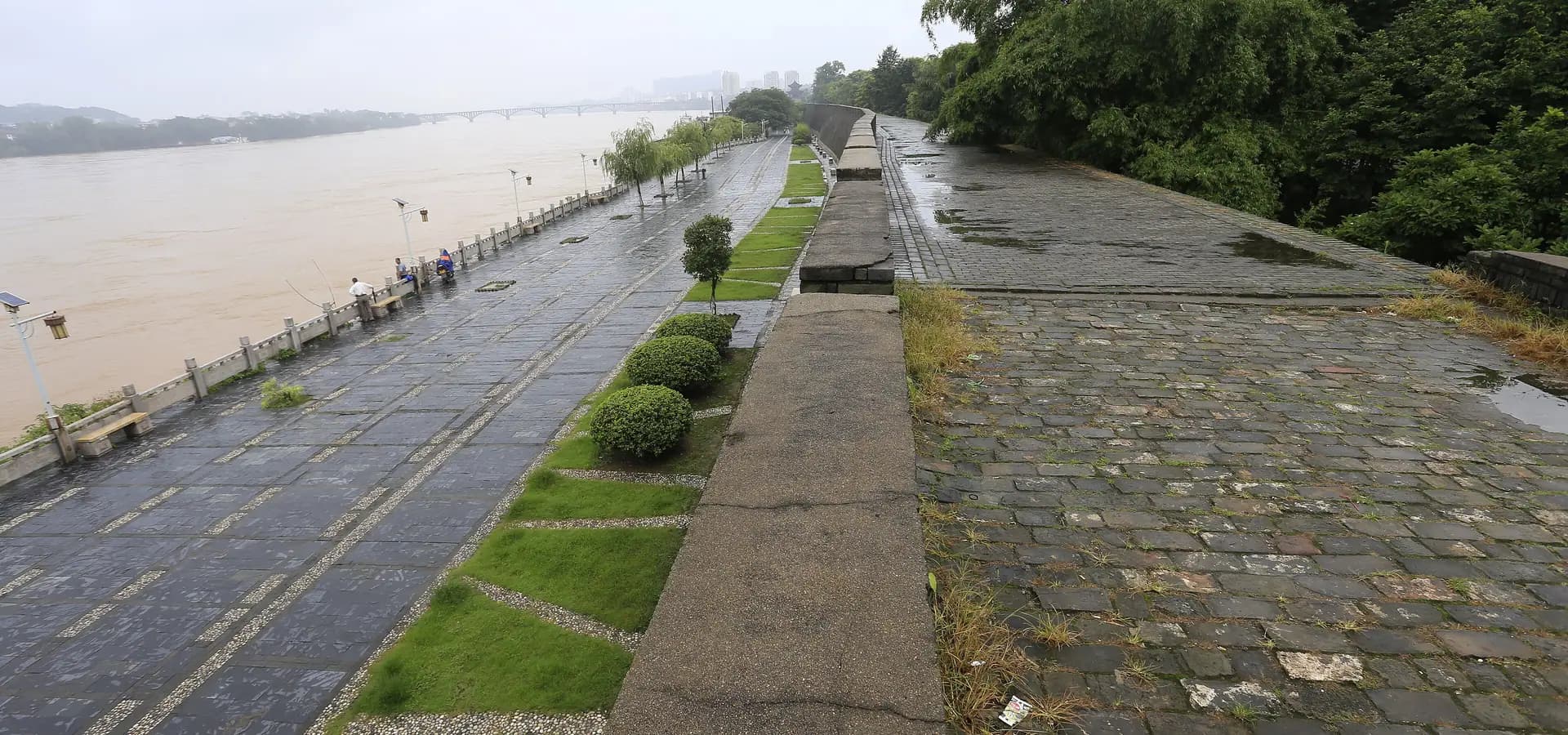 Die gepflasterte Krone der alten Stadtmauer von Ganzhou neben einem breiten Fluss, mit Bäumen, Uferweg und einer fernen Brücke