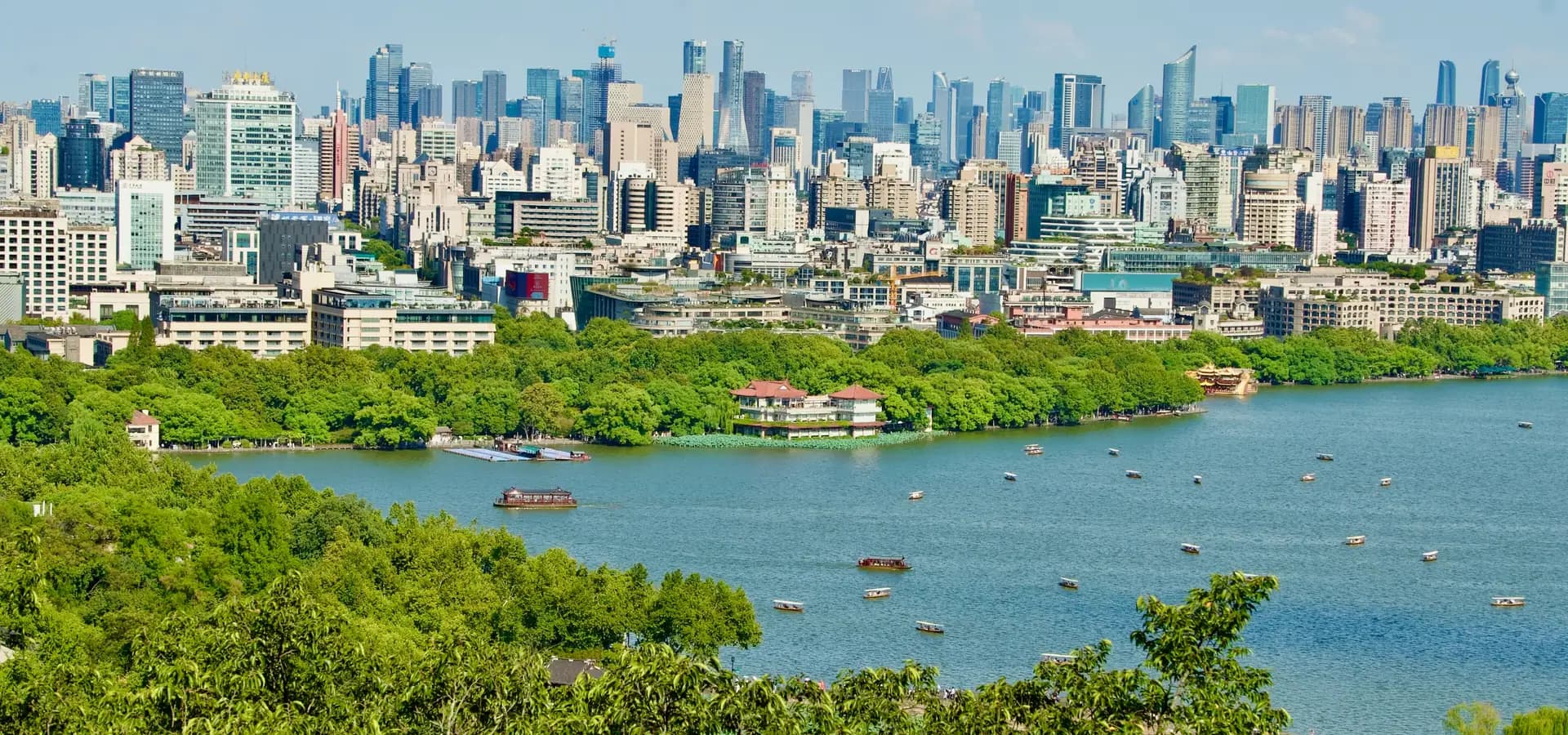 Hangzhou high-rises behind the green shore of West Lake, with small boats on the water under a blue sky
