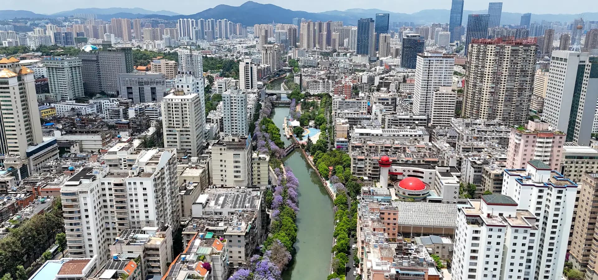 Vista aérea del centro de Kunming, un canal bordeado de jacarandás púrpuras entre rascacielos hacia colinas verdes