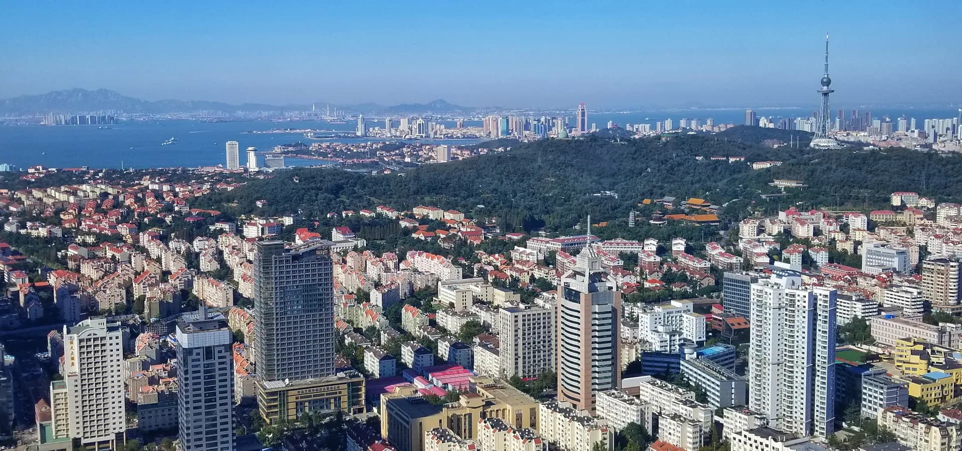 Qingdao de telhados vermelhos se espalhando abaixo da verde colina Taiping, com a baía, torres ao longe e uma torre de TV sob céu azul