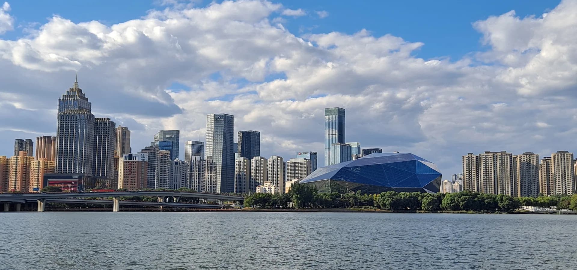 Shenyang towers and a blue faceted arts center across the wide Hun River under a cloudy blue sky
