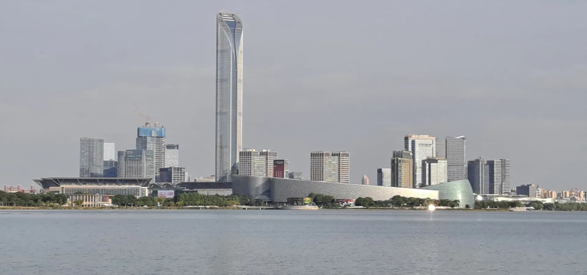 O horizonte de Suzhou à beira do lago, visto sobre as águas calmas do lago Jinji, com uma alta torre de vidro acima da margem