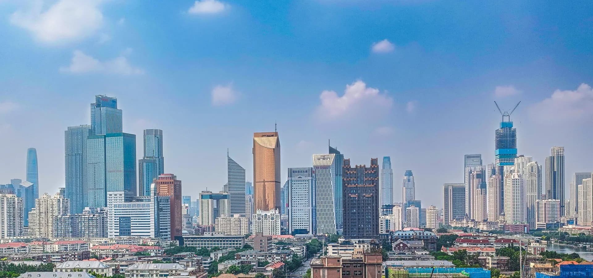 Tianjin skyline of glass towers rising behind red-roofed older buildings under a bright blue sky with clouds
