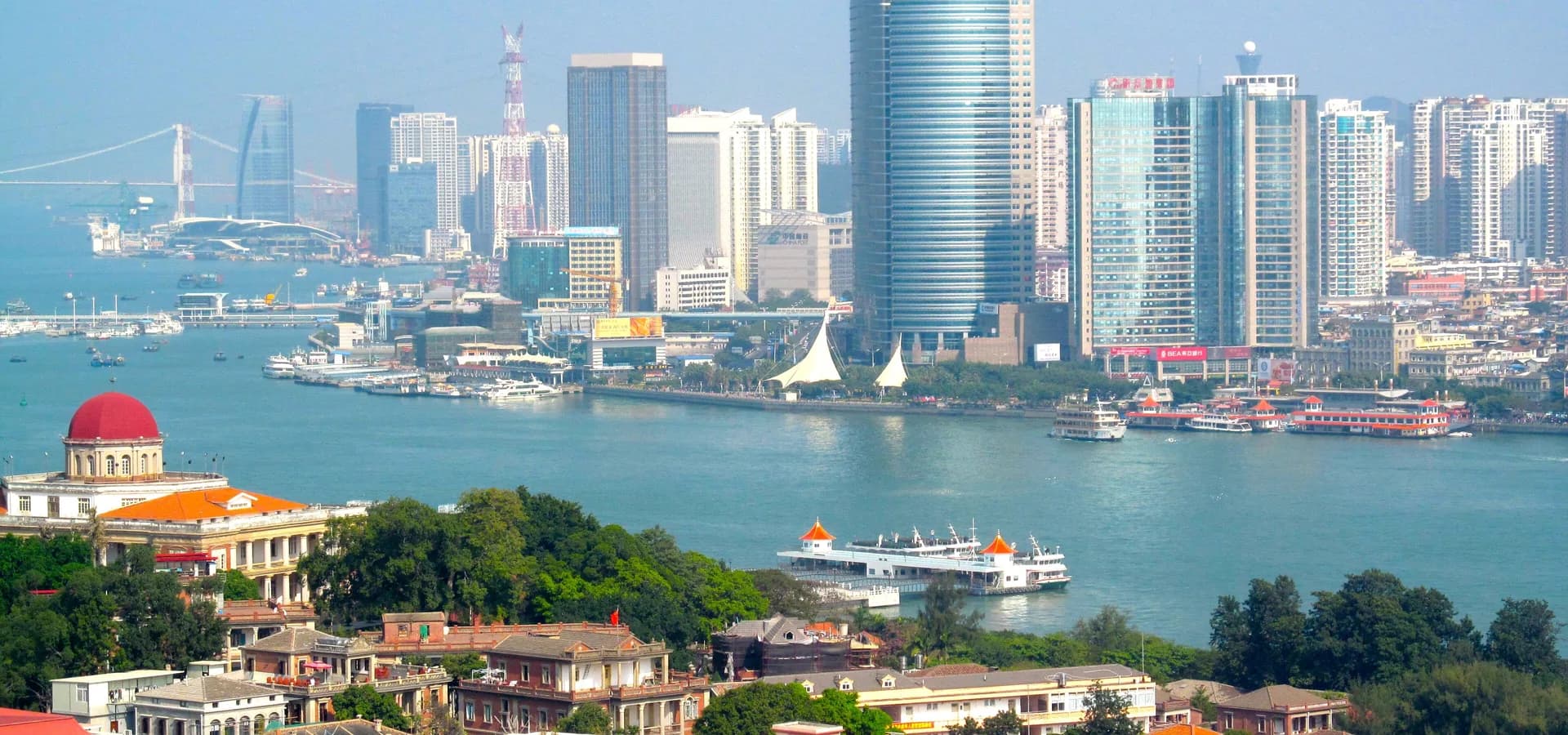 Gulangyu-Villen und eine rote Kuppel vor dem Wasser, mit Fähren und Hochhäusern von Xiamen am fernen Ufer