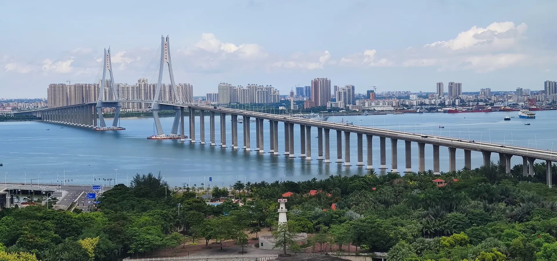Puente atirantado que cruza una amplia bahía azul hacia un horizonte de rascacielos, con un parque de palmeras al frente