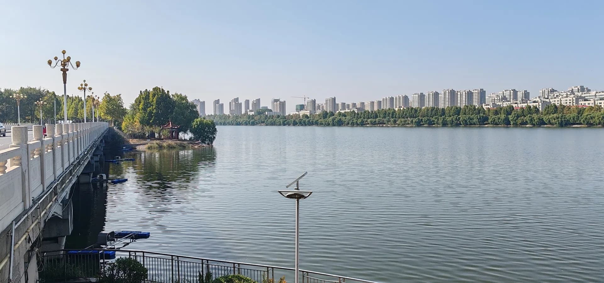 Der breite, ruhige Wei-Fluss in Zhucheng unter klarem Himmel, mit Weiden und Wohntürmen am anderen Ufer