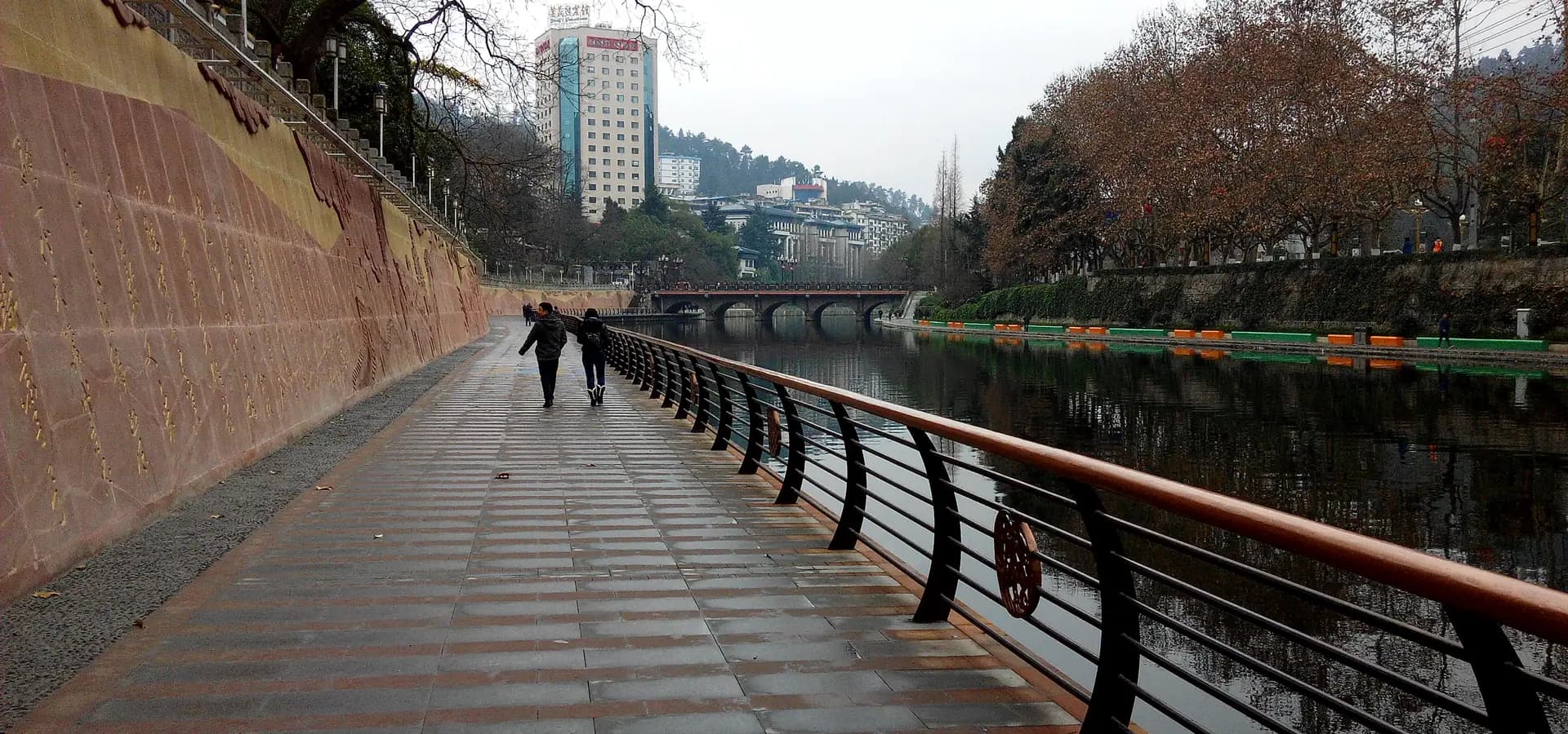 Uferweg am Fluss Xiangjiang in Zunyi mit Platanen, einer Bogenbrücke und grünen Hügeln