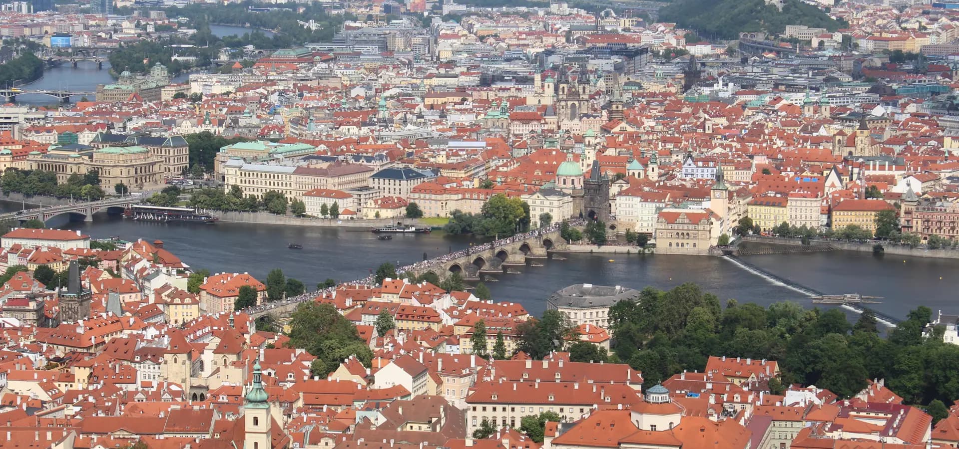 Telhados vermelhos de Praga estendendo-se até o Vltava, com a Ponte Carlos cruzando rumo às torres da Cidade Velha
