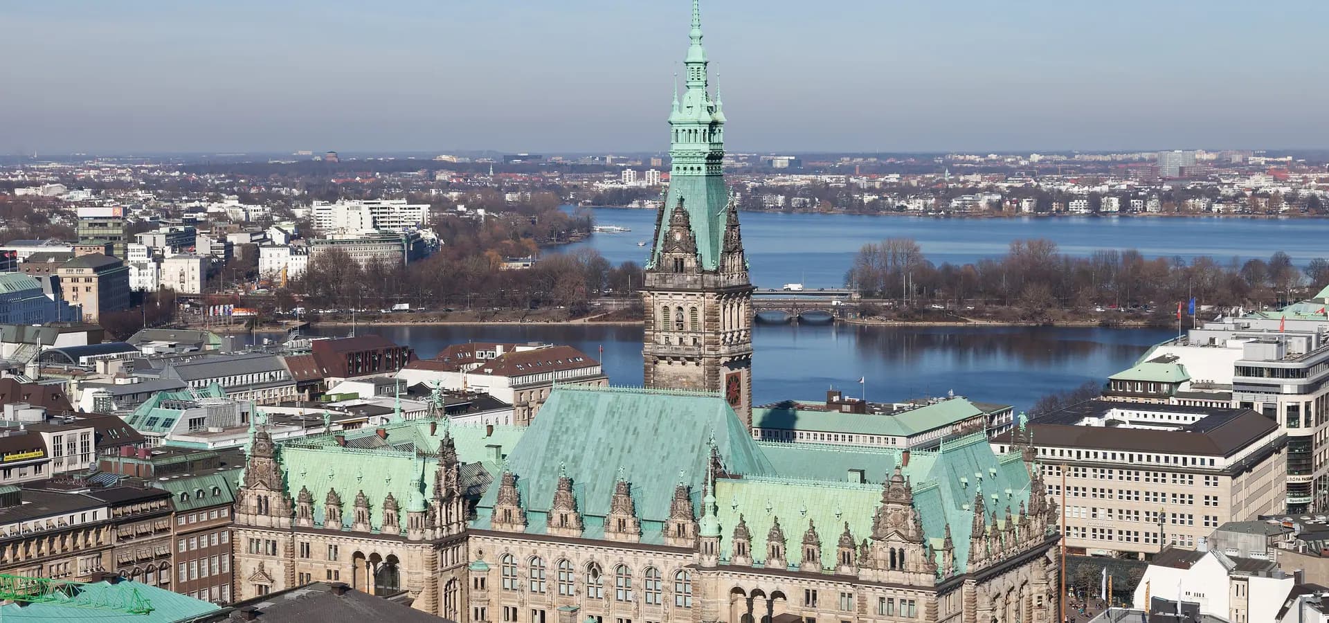 Die grünen Kupferdächer und der Turm des Hamburger Rathauses vor den blauen Alsterseen unter klarem Himmel
