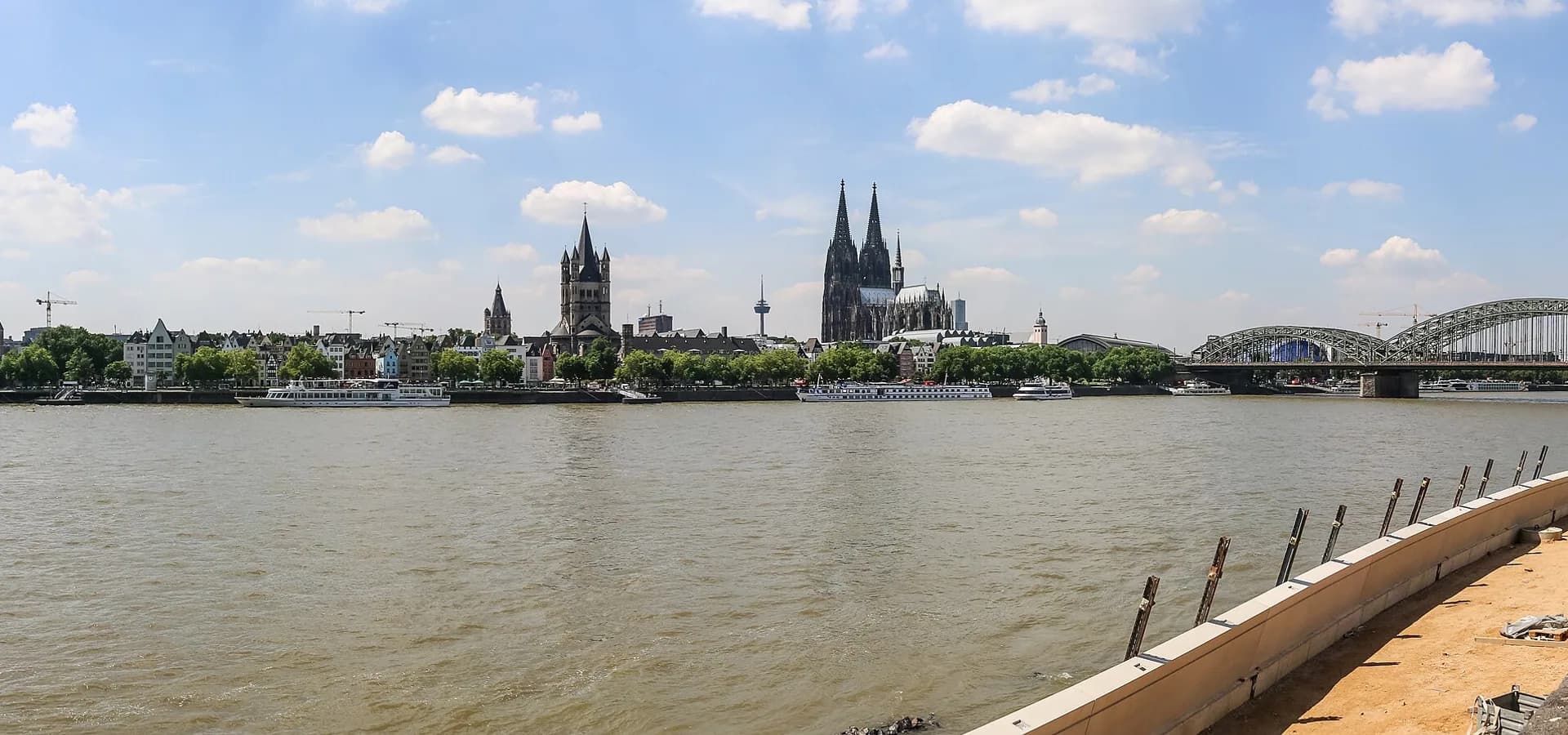 Weiter Blick über den Rhein auf die Altstadt-Skyline, die Kirche Groß St. Martin und den Kölner Dom unter blauem Himmel