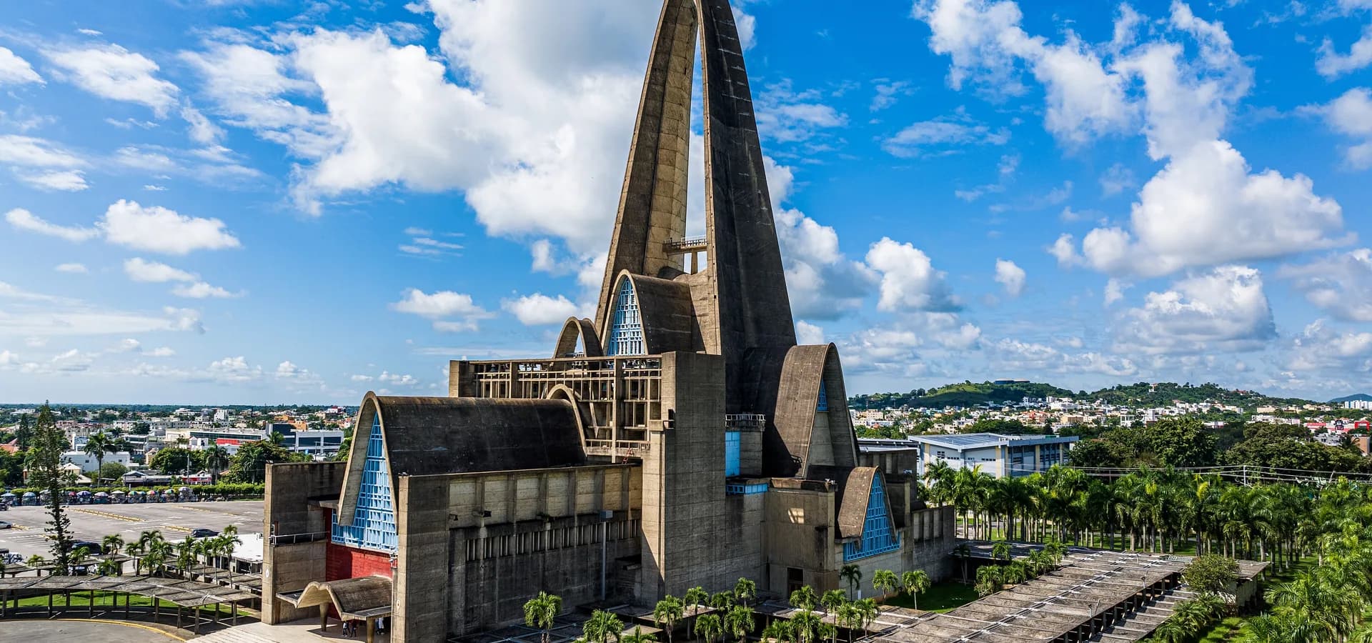 Vista aérea del arco de concreto de la basílica de Higüey sobre paseos con palmeras, con la ciudad extendiéndose hacia verdes colinas