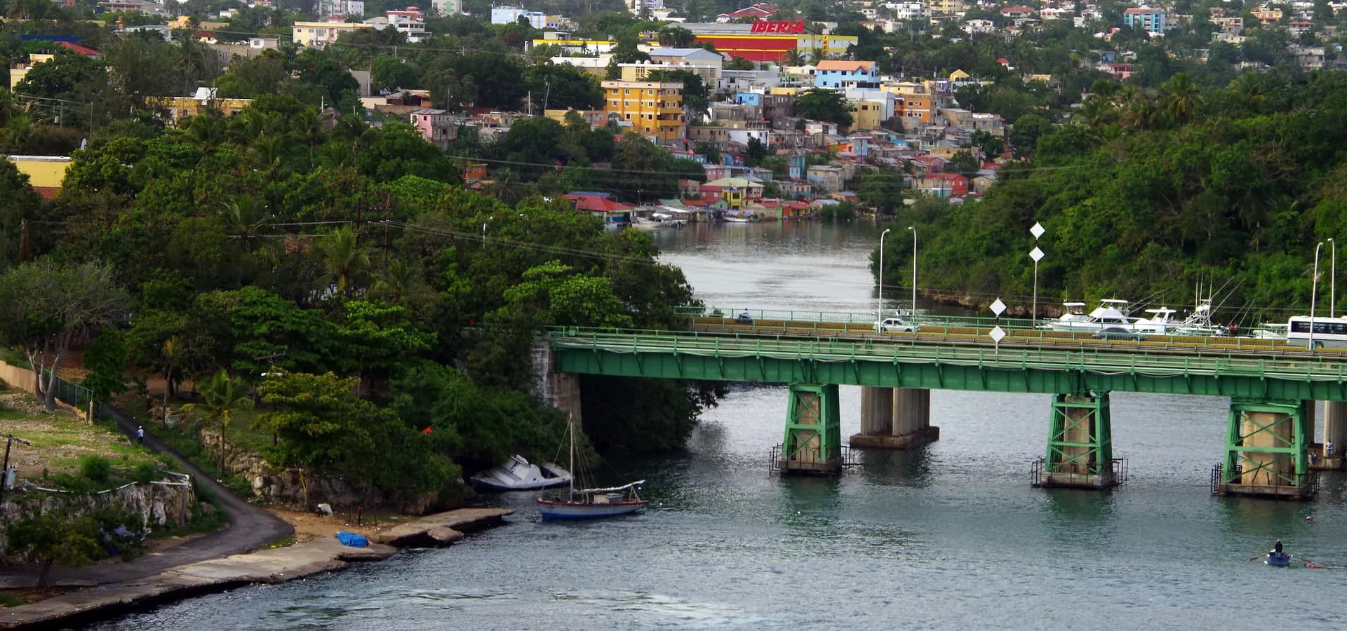 Grüne Brücke über den Río Dulce in La Romana mit Booten auf dem Wasser und bunten Häusern an den Hügeln