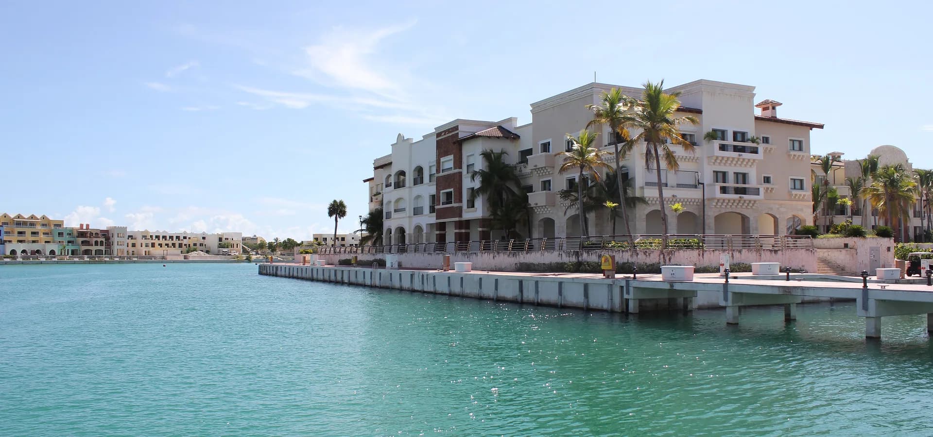Türkisfarbenes Wasser der Marina in Cap Cana mit palmengesäumten cremefarbenen Gebäuden am Kai unter blassblauem Himmel
