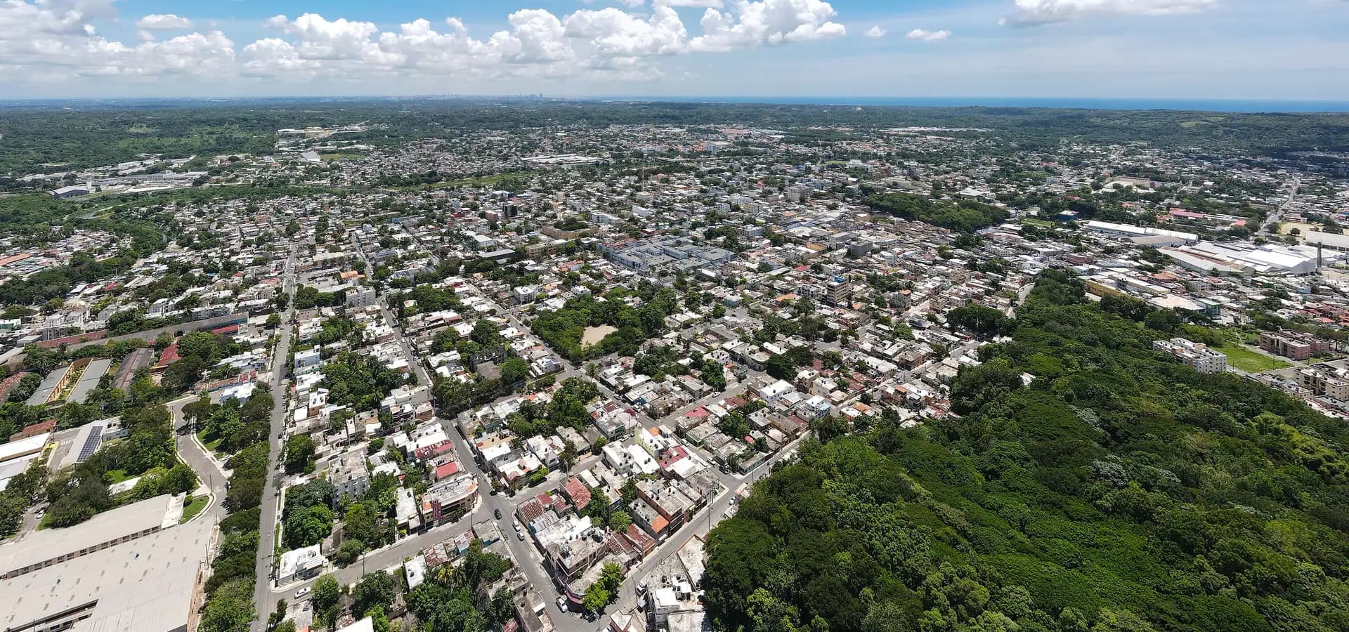 Luftbild über die Dächer von San Cristóbal und einen bewaldeten Hügel, am fernen Horizont die Karibik und Santo Domingo