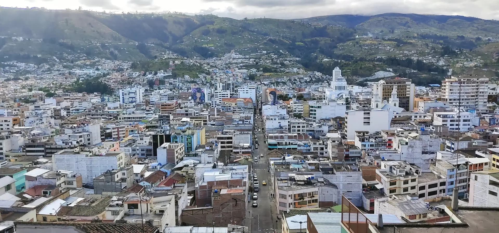 Vista panorâmica do centro de Ambato, com prédios brancos enchendo o vale sob colinas andinas verdes e um céu nublado