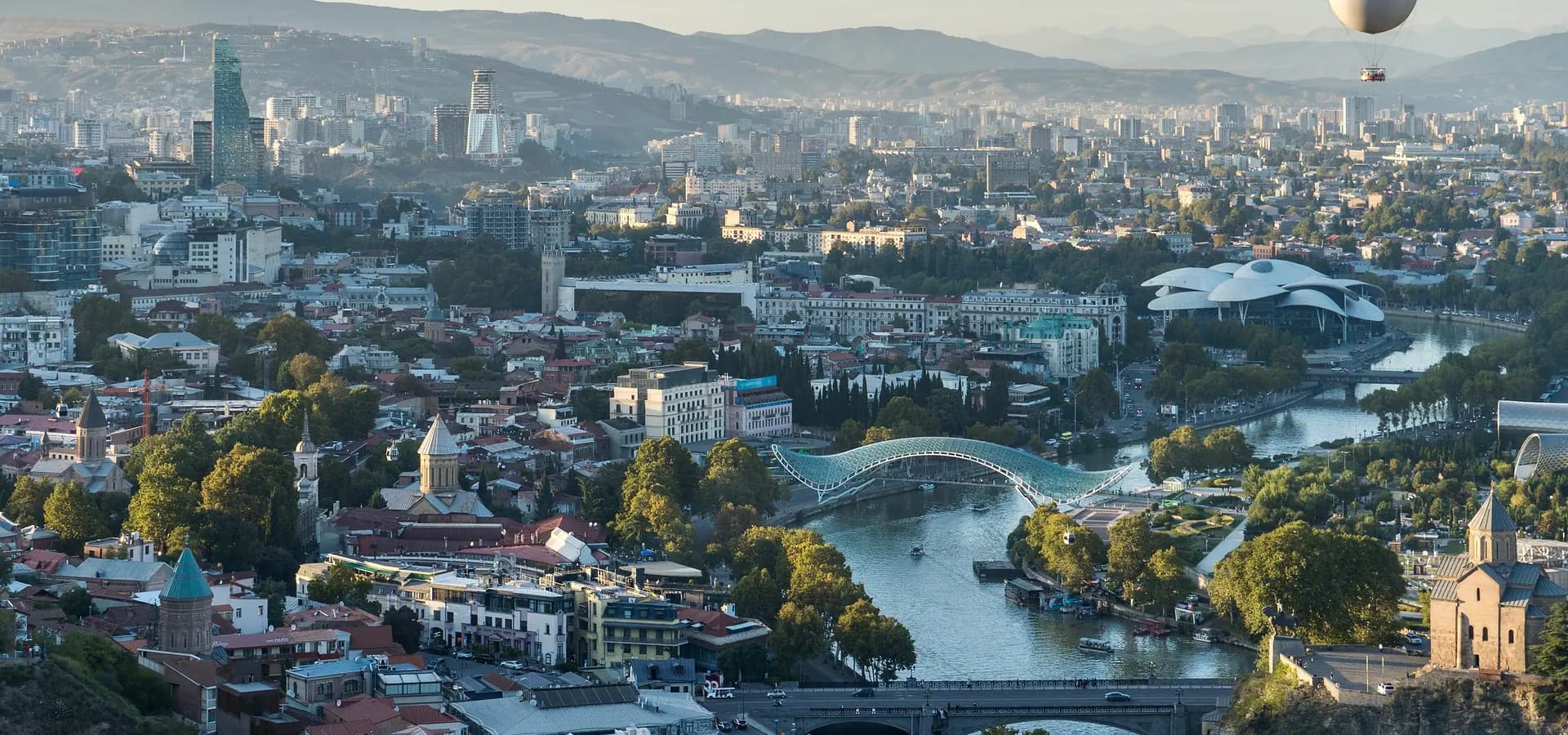 Tiflis von oben zur goldenen Stunde, die Kura schwingt sich unter der gläsernen Friedensbrücke auf ferne Hügel zu