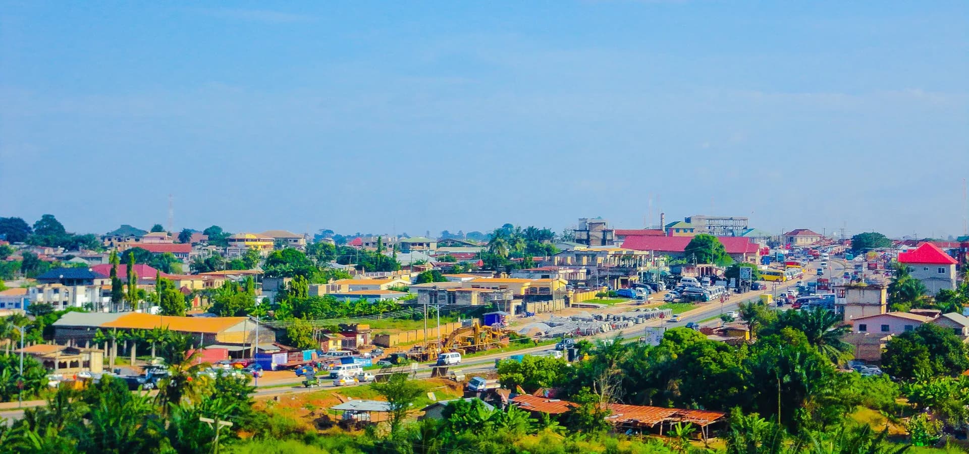 Tejados, palmeras y una avenida principal en Kronum, Kumasi, bajo un amplio cielo azul con nubes ligeras