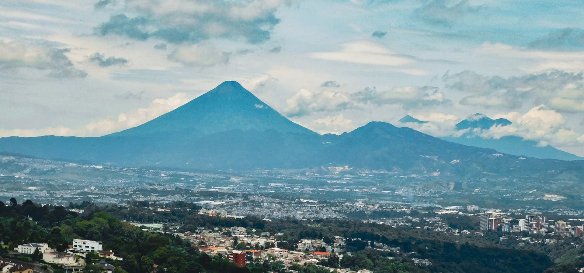 Um vulcão em forma de cone se erguendo sobre o vale da Cidade da Guatemala, com colinas arborizadas e telhados à frente