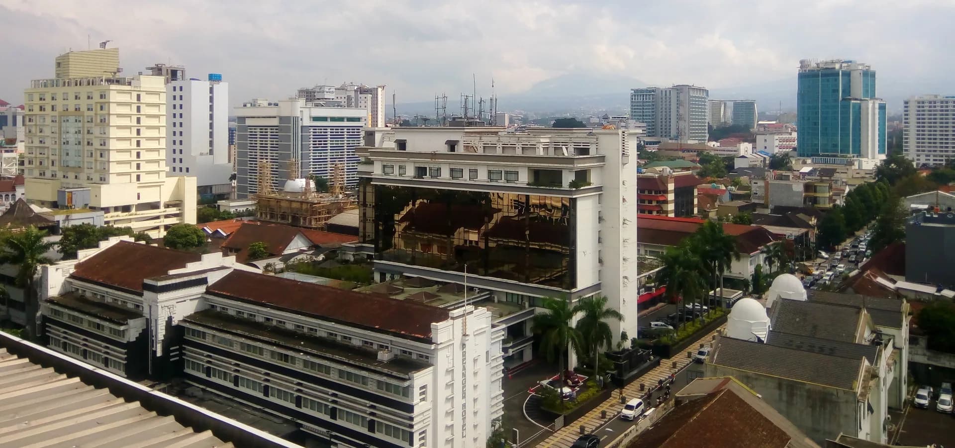 Rooftop view over central Bandung, the Art Deco Preanger hotel among towers with green hills under clouds