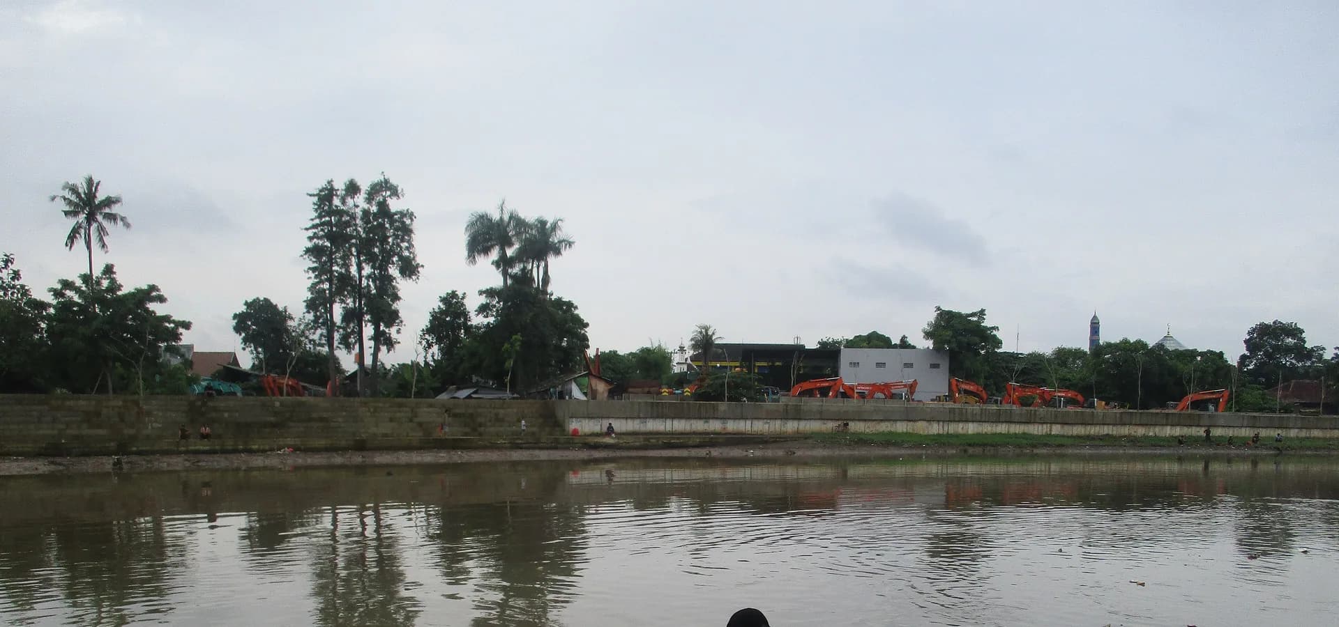 Der breite Cisadane in Tangerang unter weichem grauem Himmel, Palmen am anderen Ufer und bemalte Holzboote vertäut