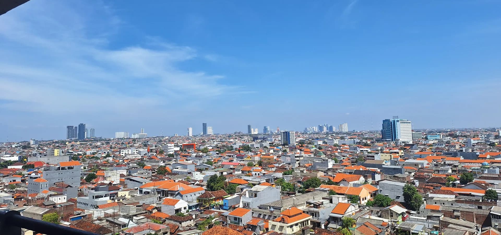 Telhados de telhas alaranjadas de Surabaya se estendendo até uma fileira de arranha-céus sob um amplo céu azul