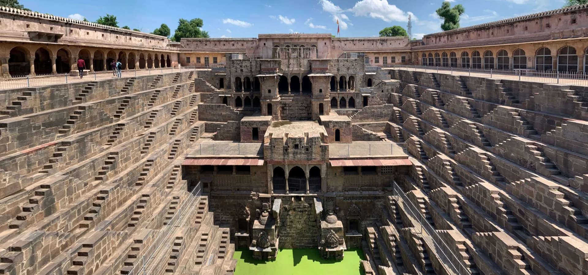 Panorama of Chand Baori, rows of stone steps on both sides leading down to green water below a carved pavilion