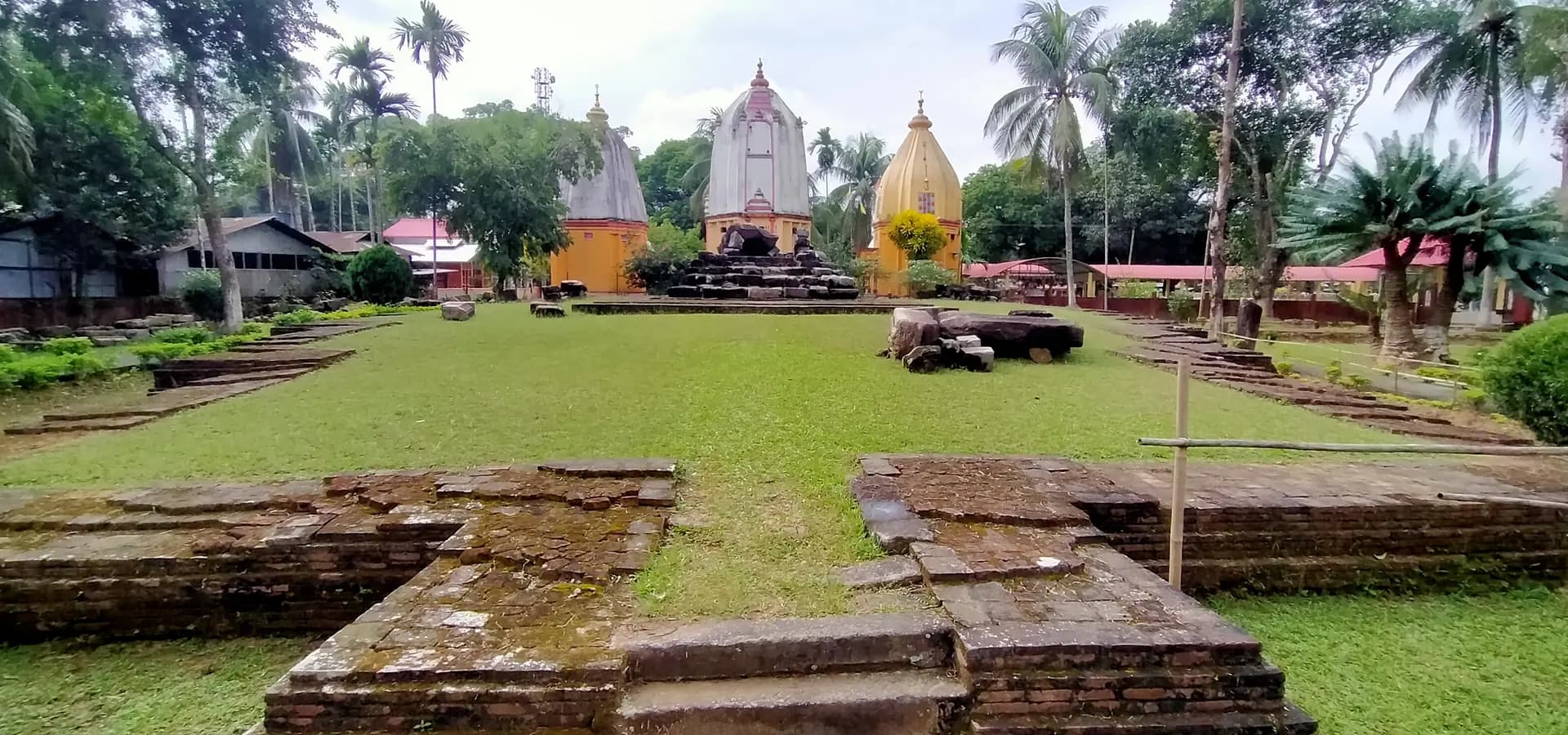 Alte Ziegeltempel-Fundamente auf grünem Rasen, dahinter Ganesh-Tempelschreine mit Kuppeln und Palmen