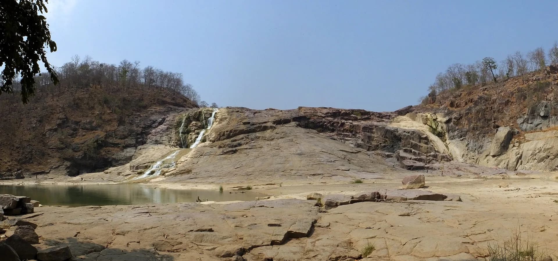 Ampla bacia rochosa da Cachoeira de Kuntala com um filete d'água, uma poça parada e colinas secas cobertas de floresta sob céu azul