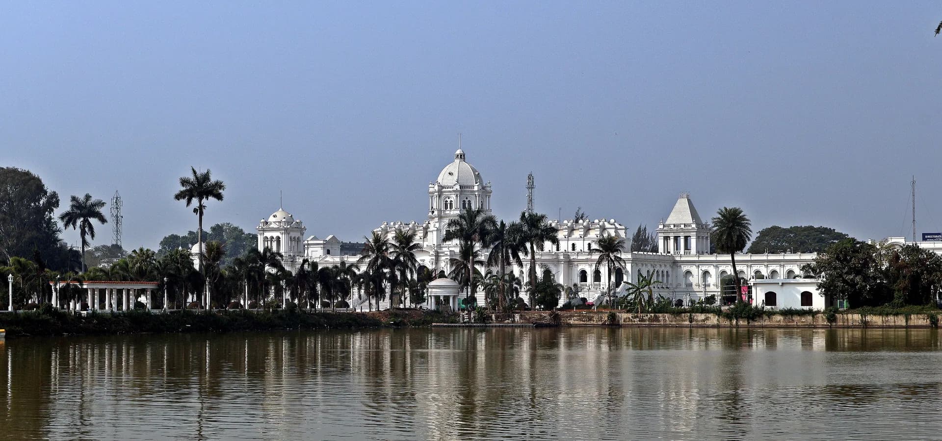 Ujjayanta-Palast und Palmen über dem ruhigen Rajbari-See unter blauem Himmel in Agartala