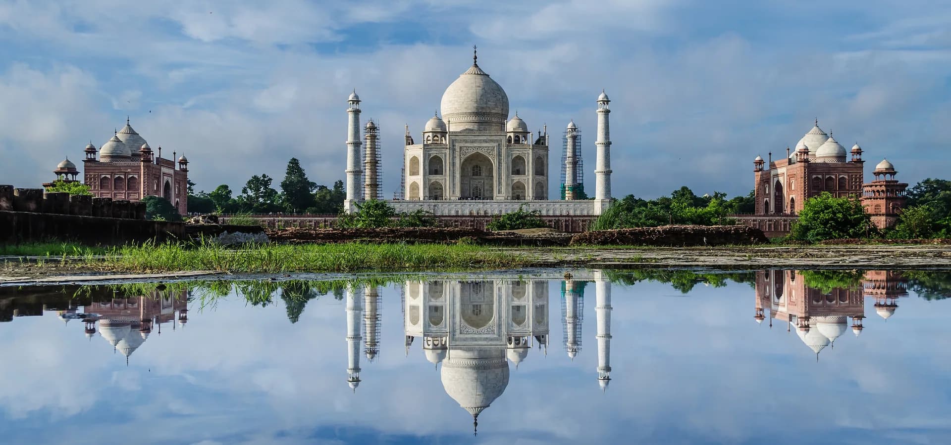 नीले आसमान और बादलों के नीचे शांत पानी में झलकता ताजमहल और उसकी लाल बलुआ पत्थर की बगल की इमारतें