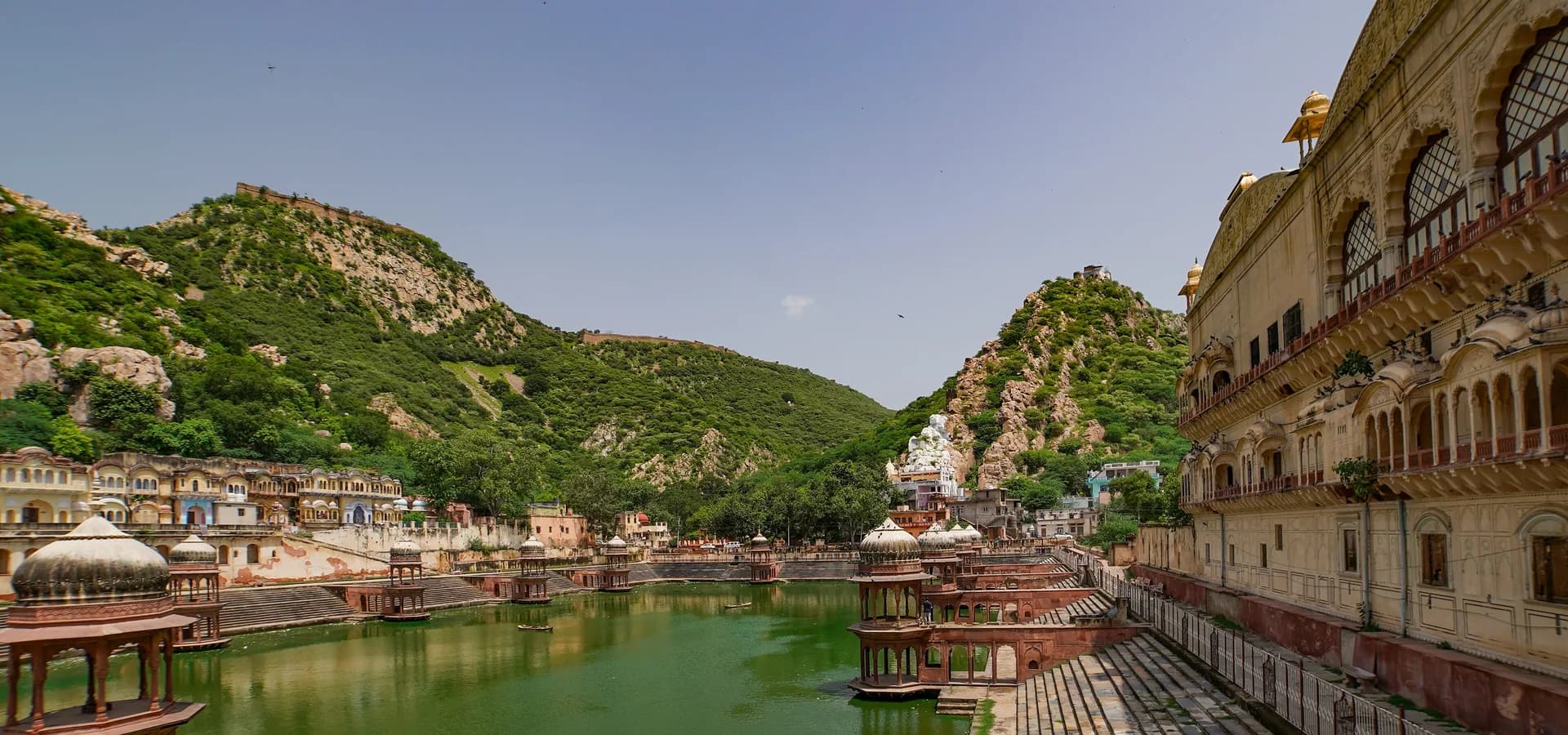 Green palace tank lined with domed pavilions and steps, palace wall on the right and green hills under a blue sky