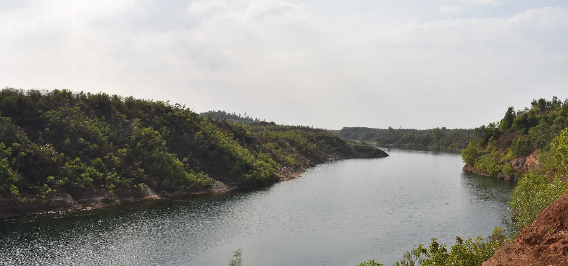 Un embalse tranquilo serpentea entre colinas verdes boscosas y orillas de tierra roja bajo un cielo nublado suave cerca de Ambikapur