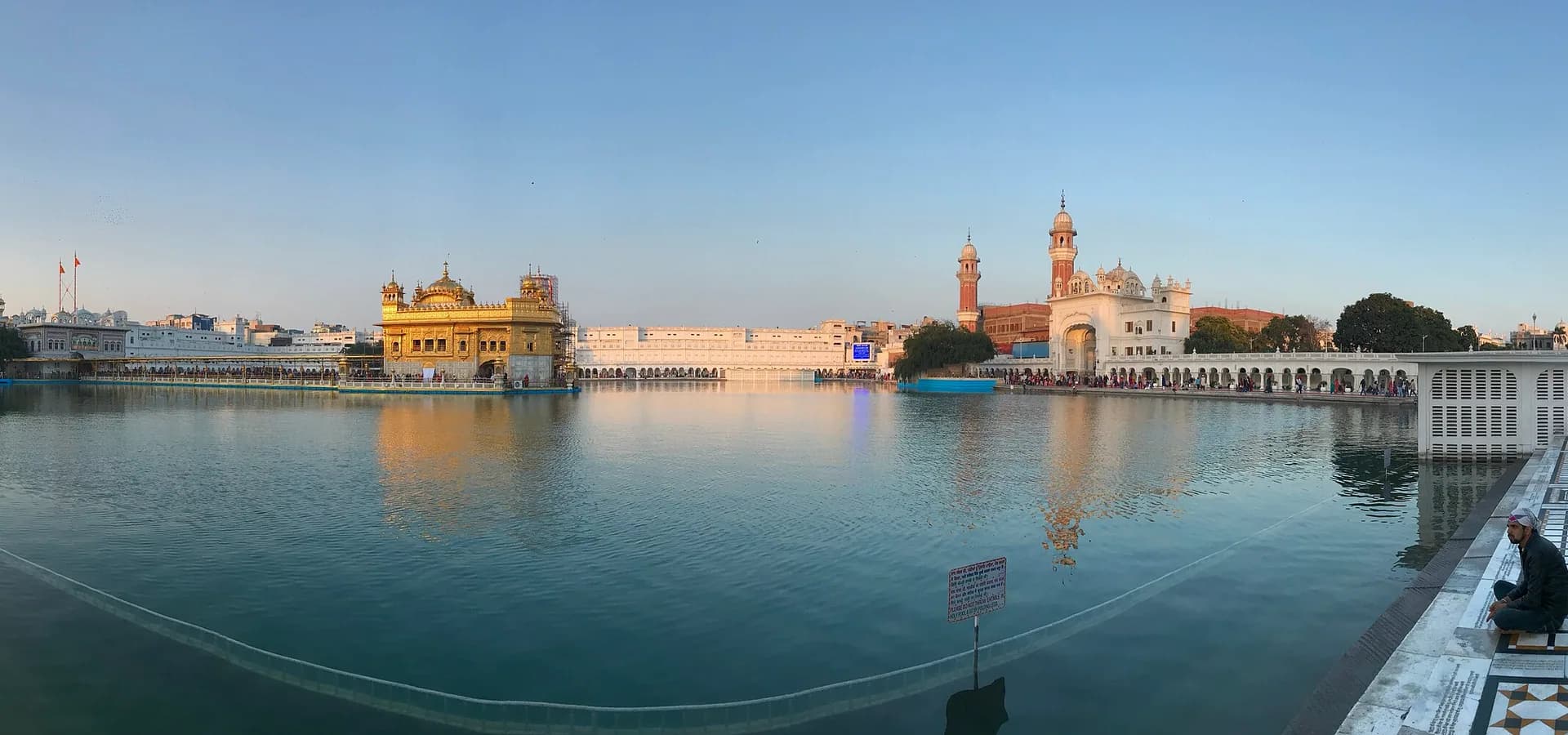 शांत पवित्र सरोवर के पार स्वर्ण मंदिर और सफ़ेद संगमरमर का परिसर, हल्के शाम के आसमान के नीचे का चौड़ा नज़ारा