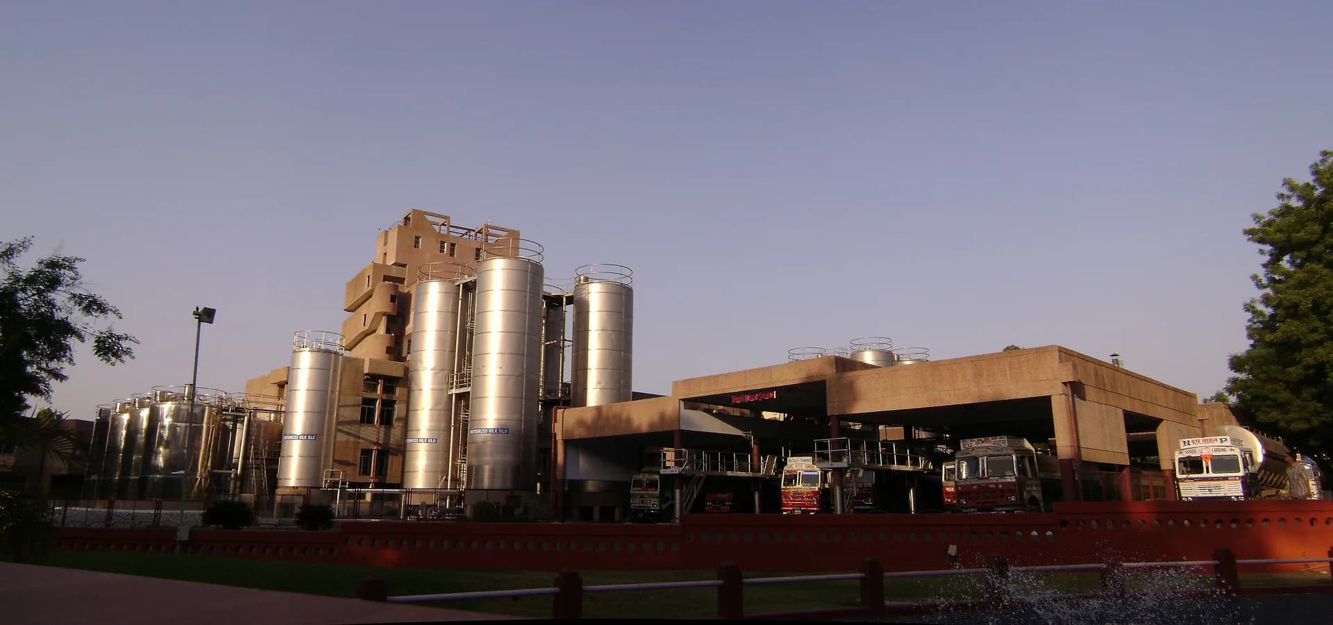 Fábrica da Amul Dairy em Anand, com altos silos de aço de leite e caminhões-tanque sob um céu azul limpo de fim de tarde