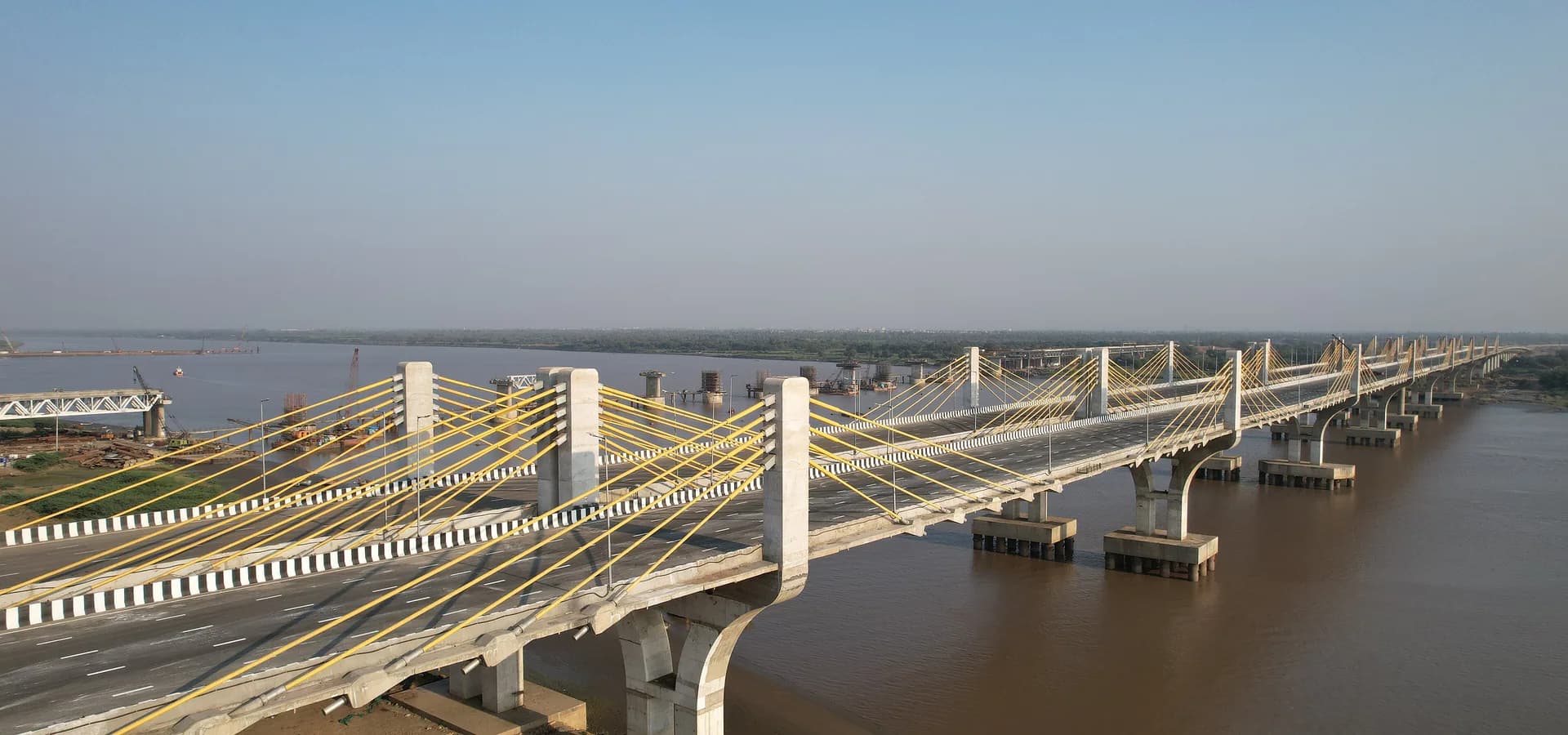 Un largo puente atirantado de cables amarillos cruza el ancho y marrón Narmada bajo un cielo azul despejado