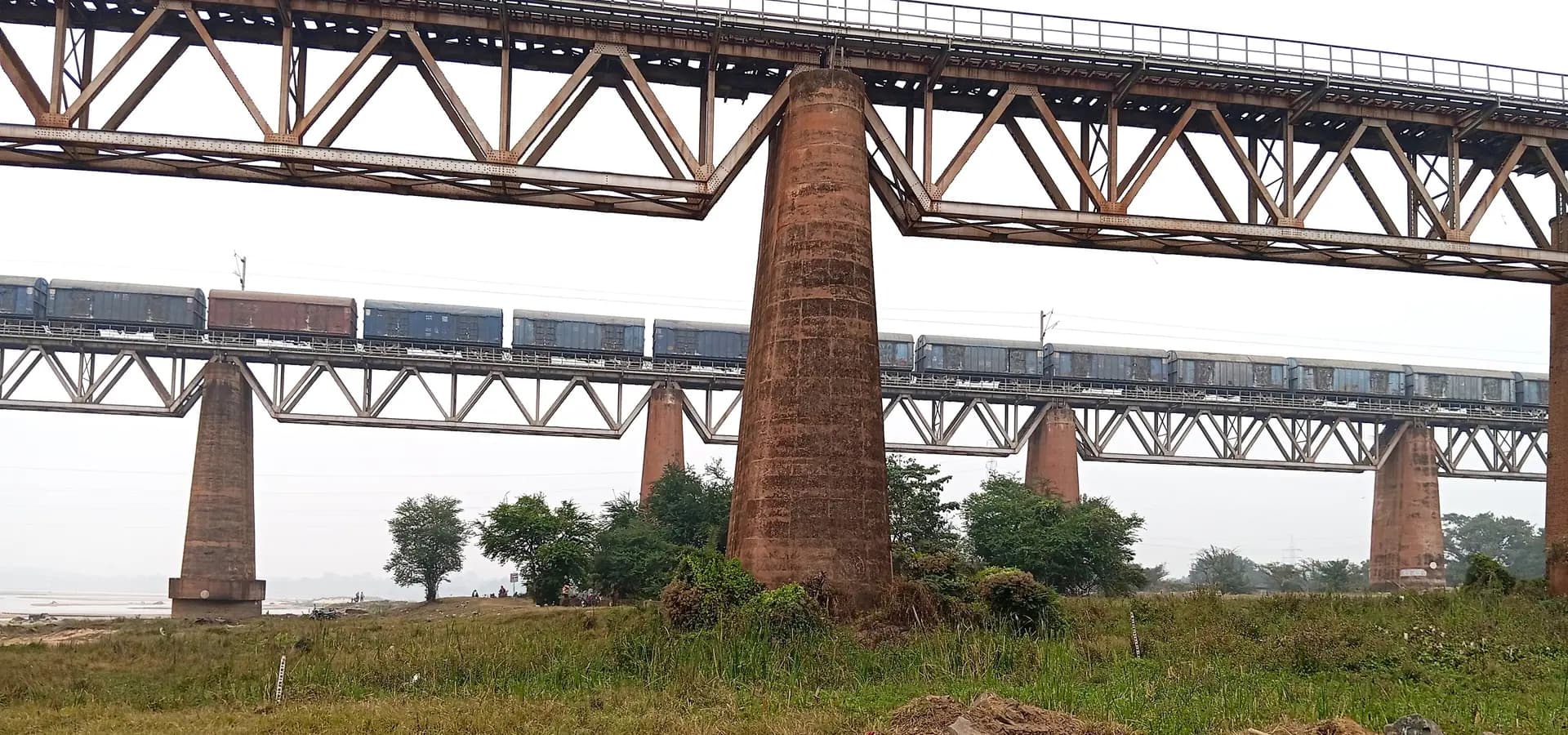 Um longo trem de carga cruzando uma ponte de treliça de aço sobre altos pilares de tijolo no rio Damodar, perto de Burnpur