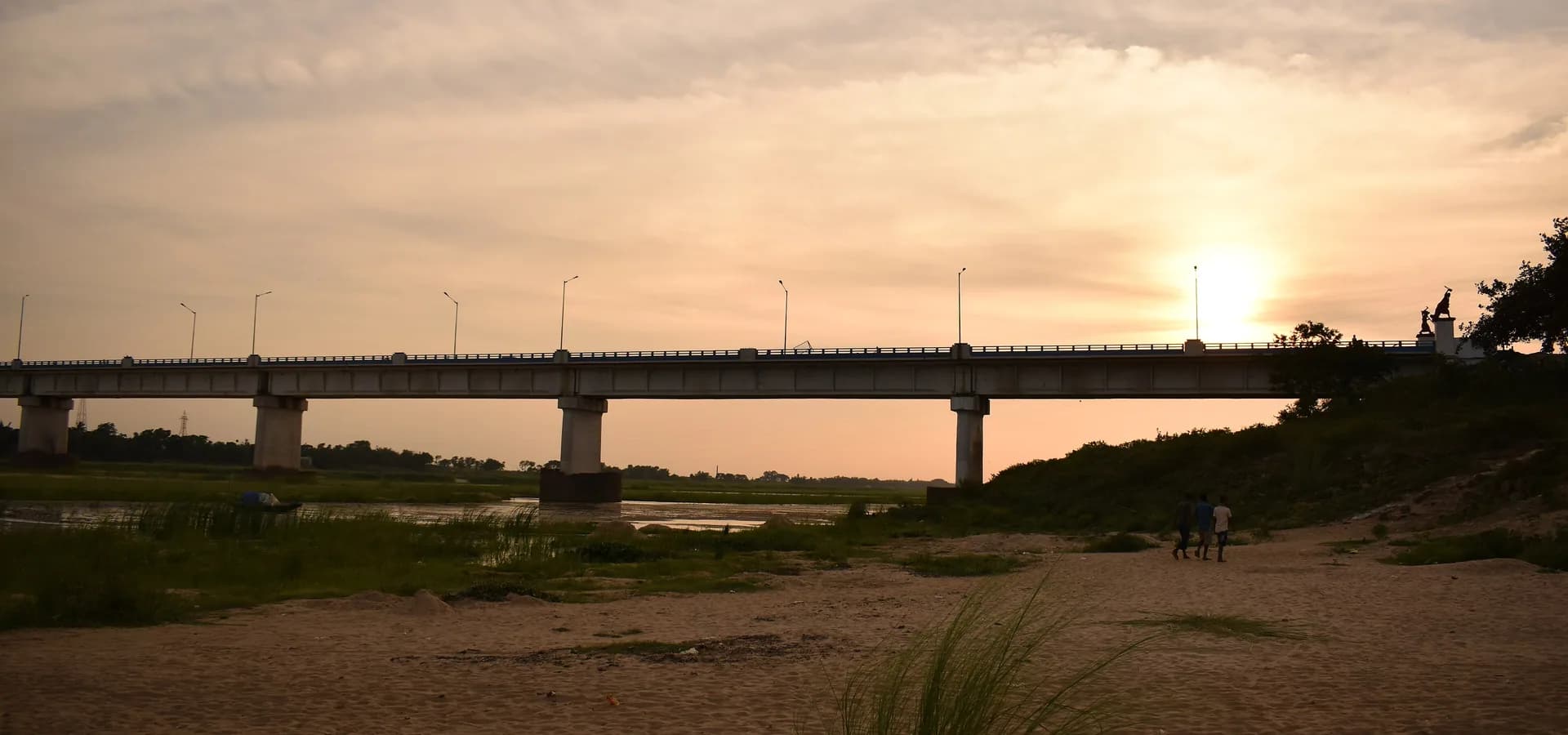 Largo puente de concreto sobre el río Damodar al atardecer, con bancos de arena y pasto en primer plano