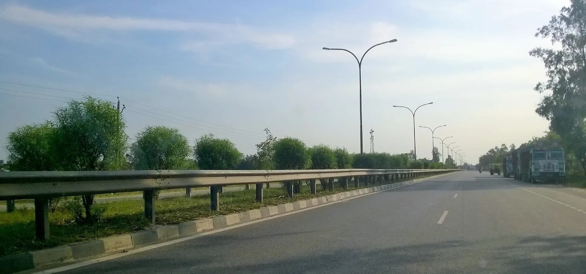 A wide highway with streetlights crossing the green, flat plain near Basti under a soft blue sky